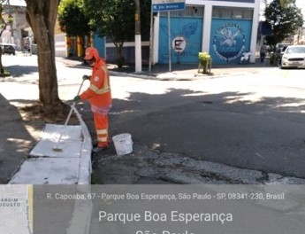 Duas tampas de concreto de boca de lobo estão sendo pintadas de branco por um funcionário da Prefeitura, com uniforme laranja. 
